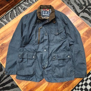 May include: A blue waxed cotton jacket featuring a brown corduroy collar, a front zip, and press-stud closures. The jacket includes four front pockets, a metal throat latch, and a Barbour brand label on the inner collar with a tartan lining.
