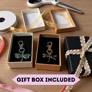 Puede incluir: Dos llaveros de lib&eacute;lula con cierres de mosquet&oacute;n se muestran en cajas de regalo de cart&oacute;n abiertas. Uno es plateado con alas de esmalte multicolor y el otro es de metal dorado. Se ven una caja negra con cinta beige, tijeras y rollos de cinta. GIFT BOX INCLUDED.
