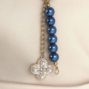 Op de afbeelding: Een goudkleurige metalen bedel met een cirkelvormige ring aan de bovenkant. De ene kant heeft een gouden ketting bevestigd aan een klaverbladvormige hanger met wit email, kleine parels en heldere kristallen. De andere kant heeft een streng van zes blauwe parels.