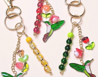 Puede incluir: Tres llaveros de metal con dijes de colibrí esmaltados, cadenas doradas y cuentas colgantes. Cada llavero incluye cuentas de vidrio o plástico en rojo, amarillo o verde, junto con pequeños dijes en forma de flor en tonos rosa y naranja.