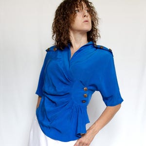 May include: A vibrant blue blouse with short sleeves and a wrap-style design. The blouse features gold-colored buttons and shoulder accents. The fabric has a slight sheen, and the style is reminiscent of vintage fashion.