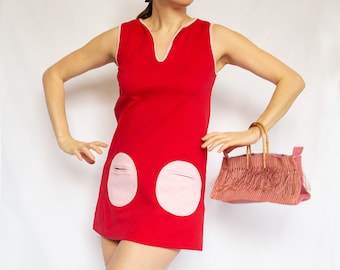 May include: A red sleeveless mini dress with pink circle pockets and a pink trim around the neckline. The dress is being worn by a person holding a pink fringed handbag with bamboo handles.