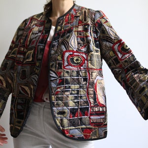 Puede incluir: Una chaqueta acolchada con un patrón geométrico en tonos de rojo, negro, verde y dorado. La chaqueta tiene un cuello alto y una cremallera.