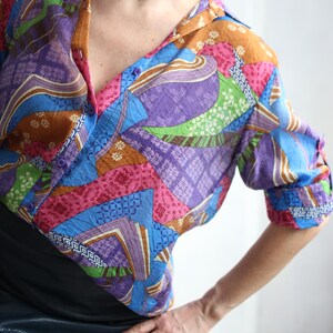 Op de afbeelding: Een kleurrijk overhemd met knopen en een patchwork-ontwerp. Het overhemd heeft een verscheidenheid aan kleuren, waaronder blauw, paars, roze, groen en bruin. De mouwen zijn opgerold en de bovenkant van het overhemd is opengeknoopt.