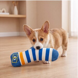 Può includere: Un cucciolo di Corgi che tiene in bocca un peluche a righe blu e bianche. Il giocattolo è cilindrico con una consistenza a coste, presenta una fascia gialla e un volto da cartone animato con occhiali quadrati ricamati in filo giallo.