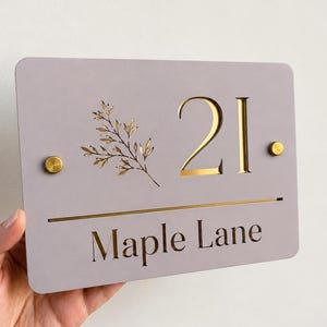 Op de afbeelding: Een rechthoekig lichtgrijs huisnummerbord met het nummer 21 en de tekst Maple Lane in gouden letters. Het bord bevat een gouden botanische takafbeelding, een horizontale gouden lijn en twee gouden ronde bevestigingsbouten aan de zijkanten.