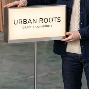 May include: A rectangular, illuminated sign with a silver metal frame mounted on a tall, thin, silver metal pole with a circular base. The sign features the black text "URBAN ROOTS" on the top line and "CRAFT & COMMUNITY" on the bottom line.