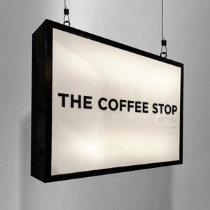 May include: A rectangular, double-sided hanging sign with a black frame and a white illuminated face. The text THE COFFEE STOP is printed in bold, black, sans-serif capital letters across the center. The sign is suspended by two thin black wires.
