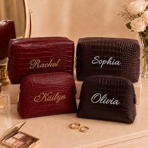 May include: Four rectangular and dome-shaped cosmetic bags with a crocodile-embossed texture. Two bags are deep red with gold lettering reading Rachel and Kailyn. Two bags are dark brown with white lettering reading Sophia and Olivia.