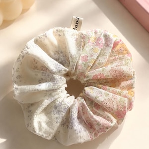 May include: A fabric hair scrunchie featuring a patchwork design of floral patterns in soft shades of pink, yellow, and blue on a white background. A small beige fabric tag with the word hand printed in black is attached to the top edge.