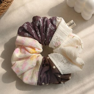 May include: A fabric hair scrunchie featuring a patchwork design of dark brown fabric with white star patterns, cream fabric with small pink floral prints, and a textured white section with lace trim. A small beige fabric tag reads land.