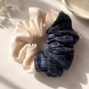 May include: A hair scrunchie featuring a two-tone design with one half made of cream-colored fabric with a subtle floral print and the other half made of dark blue fabric with a thin gold and light blue plaid pattern, including a small white tag.