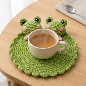 May include: A round, green crocheted coaster featuring two small, stuffed frog heads with black eyes, pink cheeks, and pink hands resting on the edge. A white ceramic mug filled with a light brown liquid sits in the center of the textured coaster.