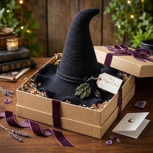 May include: A dark navy blue crocheted witch hat with a pointed crown and wide brim, nestled in a tan cardboard gift box. The hat features a green crocheted leaf accent and a tag that reads For a Magical Soul. The box is tied with a purple velvet ribbon.