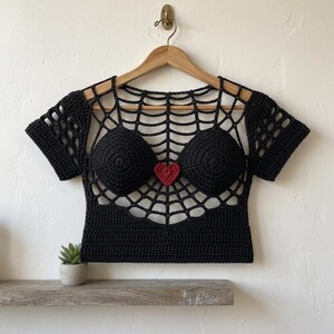Op de afbeelding: Een zwarte gehaakte crop top met een spinnenwebpatroon over de borst en schouders, korte mouwen en twee geïntegreerde ronde cups. Een klein, effen rood gehaakt hartje is in het midden tussen de twee cups op het webpatroon geplaatst.