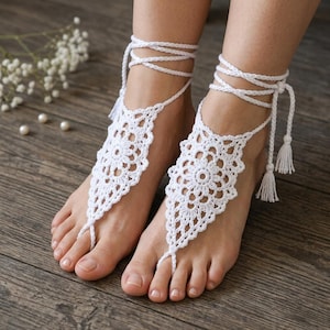 May include: A pair of white crochet barefoot sandals featuring an intricate floral mandala pattern that covers the top of the foot. Each sandal includes a loop for the second toe and long braided ties that wrap around the ankle, finished with tassels.