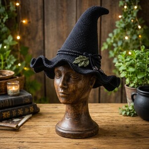 May include: A dark navy blue crocheted witch hat with a tall, pointed crown and a wide, wavy brim. The hat features a decorative band with a small, textured green leaf accent and dark beads tied to the side, resting on a wooden mannequin head.
