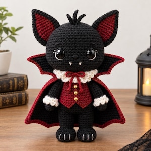 May include: A black crocheted bat doll with large dark eyes and two white fangs. It wears a red vest with three gold buttons, a white ruffled collar with a red bow, and a black cape with a red lining. The doll has pointed ears with red centers.