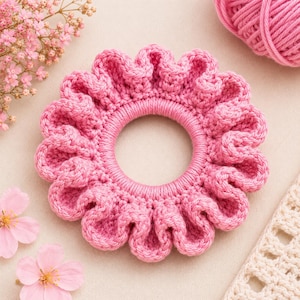 May include: A bright pink crocheted hair scrunchie with a ruffled, circular design. The accessory features a dense, textured yarn construction with a central elastic opening, showcasing a consistent, vibrant pink color throughout the entire piece.