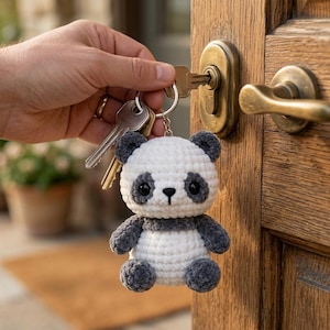 Pode incluir: Um porta-chaves de panda em croché com corpo de fio branco e cinzento escuro, orelhas redondas, olhos de segurança pretos e um pequeno nariz bordado. O panda de pelúcia está preso a um anel de metal prateado com duas chaves de casa.