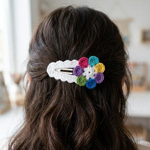 May include: A hair clip featuring a white crocheted base with a scalloped edge and a multi-colored crocheted flower attached to the side. The flower petals are pink, light blue, yellow, purple, dark blue, and green, surrounding a white center.