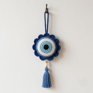 May include: A crocheted wall hanging featuring a circular evil eye motif with concentric rings of black, light blue, white, and dark blue. The piece has a scalloped dark blue border, a braided hanging loop, and a blue tassel with gold and white beads.