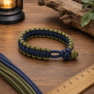 Peut inclure: Un bracelet en paracorde avec un motif tressé en vert olive et bleu marine. Le bracelet comprend un mécanisme de fermeture à boucle et nœud. Une règle en bois est visible, indiquant que le bracelet mesure environ 19 cm de longueur.