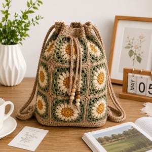 May include: A drawstring backpack constructed from crocheted granny squares featuring a daisy motif in yellow, white, and sage green. The bag includes tan yarn straps and two dangling wooden beads at the end of the drawstring closure cords.
