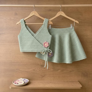 May include: A sage green crochet two-piece set consisting of a sleeveless wrap-style crop top with a side tie and a matching flared skirt. The top features a small pink crocheted flower accent. A small bowl with additional crochet flowers sits below.