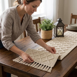 May include: A cream-colored rectangular crochet table runner featuring a raised diamond pattern and decorative fringe at both ends. The runner is crafted from thick yarn and displays a textured, geometric design that spans the length of the fabric.