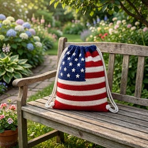 May include: A drawstring backpack featuring a crocheted design of the American flag with a blue canton containing white stars and horizontal red and white stripes. The bag includes thick white rope cords for closure and carrying, showcasing a textured yarn construction throughout the entire piece.