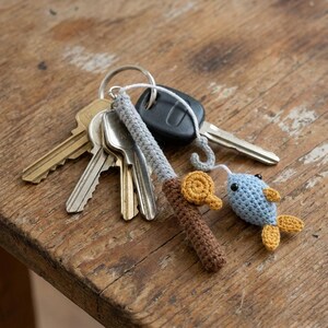 May include: A crochet keychain featuring a miniature fishing rod with a brown handle, a grey shaft, and a small yellow reel. A white string connects the rod to a light blue fish with yellow fins and black bead eyes, attached to a metal ring with keys.