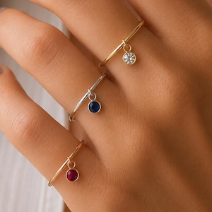 May include: Three thin metal rings worn on fingers, each featuring a small, dangling circular charm. The top ring is gold with a clear stone, the middle is silver with a blue stone, and the bottom is gold with a deep red stone.