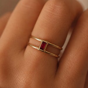 May include: A gold ring featuring a double band design connected by a single, vertically oriented, rectangular baguette-cut red gemstone set in the center. The metal has a polished finish, and the stone is held in a simple, clean bezel setting.