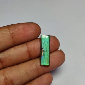 Puede incluir: Una piedra preciosa de labradorita rectangular con una superficie pulida de iridiscencia verde y azul vibrante. La piedra muestra fracturas internas naturales y un juego de colores distintivo en su cara plana, con un acabado liso y brillante.