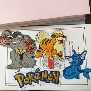 May include: Four embroidered patches featuring three cartoon creatures and a yellow and blue Pokémon logo. One patch shows a grey muscular figure with red accents, another depicts an orange and black striped creature, and one shows a blue aquatic creature.