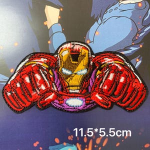 May include: An embroidered patch featuring a red and gold armoured character with glowing blue eyes and a chest piece, posed with both fists forward. The patch measures 11.5 by 5.5 centimetres and has a black border with detailed stitching.