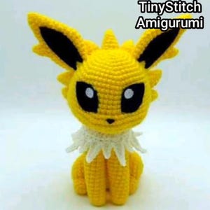 May include: A yellow crocheted amigurumi creature with large pointed ears featuring black inner sections, black eyes with white pupils, and a white ruffled collar around the neck. The text TinyStitch Amigurumi is visible in the upper right corner.