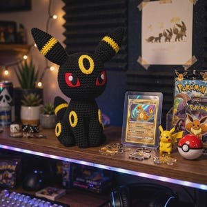 Könnte beinhalten: Ein gehäkeltes Umbreon-Plüschtier in Schwarz und Gelb mit roten Augen, spitzen Ohren mit gelben Ringen sowie gelben ovalen Markierungen auf Stirn und Körper, das aufrecht auf einem Schreibtisch aus Holz neben verschiedenen Gaming-Sammlerstücken sitzt.