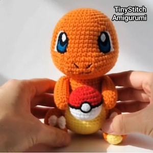 Op de afbeelding: Een gehaakt amigurumi-figuur van oranje garen met grote blauw-witte ogen en een gele buik. Het figuur houdt een kleine gehaakte bal in rood, wit en zwart vast. De tekst TinyStitch Amigurumi is zichtbaar in de rechterbovenhoek van de afbeelding.