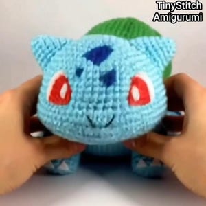 May include: A light blue crocheted amigurumi creature with a large green bulb on its back, red eyes, and dark blue forehead markings. The text TinyStitch Amigurumi is visible in the top right corner of this handmade yarn plush toy.