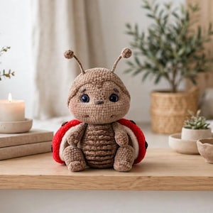Pode incluir: Um brinquedo de joaninha feito à mão em crochê com cabeça e corpo castanho-claros e costuras texturizadas em castanho-escuro. O brinquedo tem duas antenas no topo da cabeça, olhos grandes e escuros e asas vermelho-brilhantes com pintas pretas.