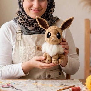 May include: A crocheted Eevee plush toy featuring a light brown yarn body, large pointed ears with dark brown inner sections, black and white eyes, and a fluffy white yarn collar around the neck. The figure is seated in a stable upright position.