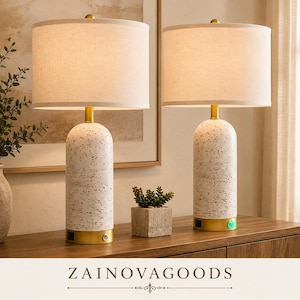 May include: A pair of table lamps featuring cylindrical white bases with a speckled texture, topped with off-white fabric drum shades. Each lamp has a gold-toned metal base with a built-in power outlet and a circular power button on the front.