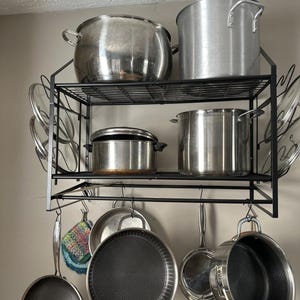 May include: A black metal wall-mounted pot rack with two wire shelves and a bottom hanging bar. The rack holds various stainless steel pots, pans, and lids. Several pans with textured interiors hang from hooks on the bottom bar of the rack.