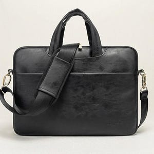 Könnte beinhalten: Eine schwarze, rechteckige Laptoptasche mit glatter Oberfläche, ausgestattet mit zwei Tragegriffen und einem abnehmbaren Schultergurt mit Polsterung. Die Tasche verfügt über eine Vordertasche, silberfarbene Metallbeschläge und ein kleines Logo.