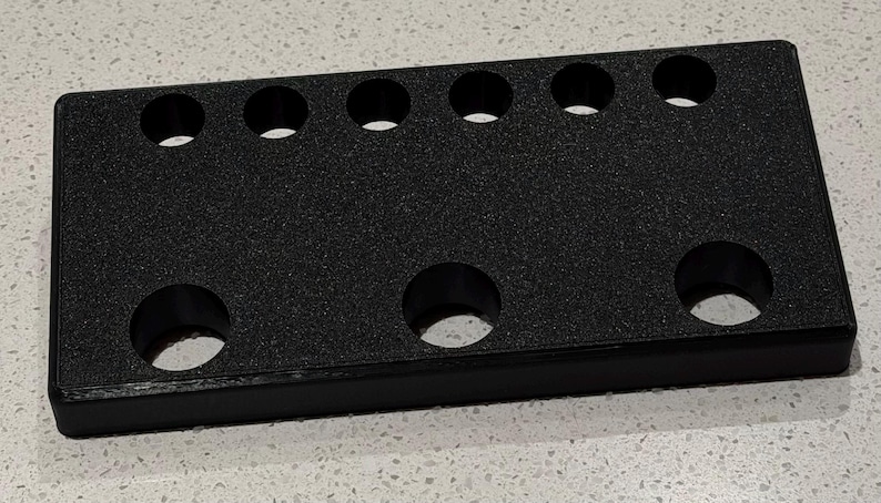 May include: A rectangular black organizer tray with a textured surface featuring six small circular holes arranged in a single row along the top edge and three larger circular holes positioned in a row along the bottom edge of the unit.