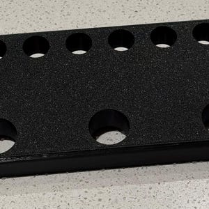 Puede incluir: Una bandeja organizadora rectangular de color negro con superficie texturizada, que presenta seis orificios circulares peque&ntilde;os dispuestos en una fila en el borde superior y tres orificios circulares m&aacute;s grandes en el borde inferior.