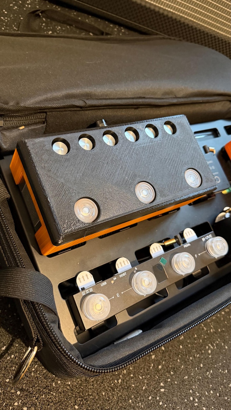 Puede incluir: Un controlador electr&oacute;nico rectangular negro y naranja con seis botones circulares plateados en la parte superior, colocado sobre una pedalera negra con m&uacute;ltiples interruptores circulares transparentes, etiquetas como USB y HOST, y el texto M-VAVE.