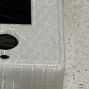 May include: A translucent, 3D-printed rectangular object with a textured surface finish. The piece features a large square cutout in the center and two circular holes positioned along the side, showing visible layer lines from the manufacturing process.